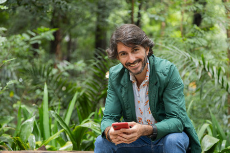 View of a young man with smartphone at daytime sitting down on a bench in a green park in the city. High quality photo.の写真素材