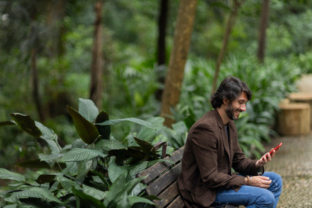 View of a young man with smartphone at daytime sitting down on a bench in a green park in the city. High quality photo.の写真素材