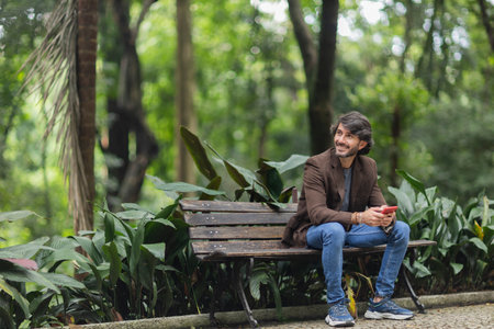 View of a young man with smartphone at daytime sitting down on a bench in a green park in the city. High quality photo.の写真素材