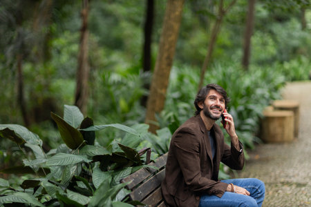 View of a young man with smartphone at daytime sitting down on a bench in a green park in the city. High quality photo.の写真素材