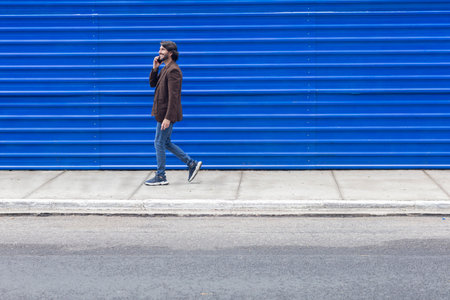 View of a young man with smartphone walking on the street at daytime with a blue background. High quality photo.の写真素材