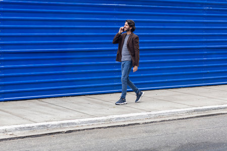 View of a young man with smartphone walking on the street at daytime with a blue background. High quality photo.の写真素材