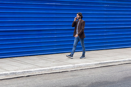 View of a young man with smartphone walking on the street at daytime with a blue background. High quality photo.の写真素材