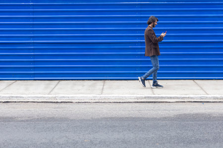 View of a young man with smartphone walking on the street at daytime with a blue background. High quality photo.の写真素材