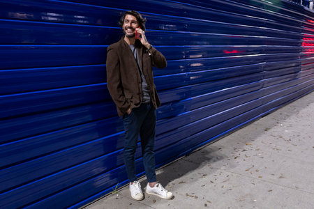 Young man using a smartphone at night time leaning against a blue wall with city lights. Young man using a smartphone at night time leaning against a blue wall with city view landscape in the background.の写真素材