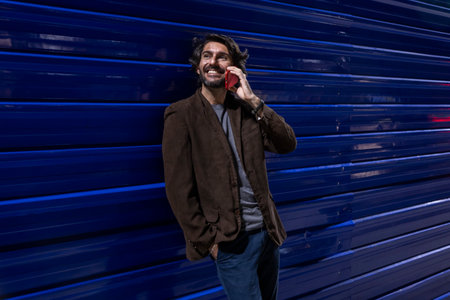 Young man using a smartphone at night time leaning against a blue wall with city lights. Young man using a smartphone at night time leaning against a blue wall with city view landscape in the background.の写真素材