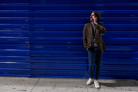 Young man using a smartphone at night time leaning against a blue wall with city lights. Young man using a smartphone at night time leaning against a blue wall with city view landscape in the background.の写真素材
