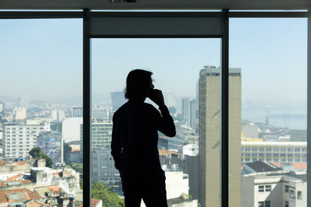 Silhouette of a young man speaking on mobile phone in the office looking outside with city view in the background.の写真素材