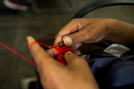Close-up of hands knitting with red yarn using a crochet hook. The image highlights craftsmanship,...