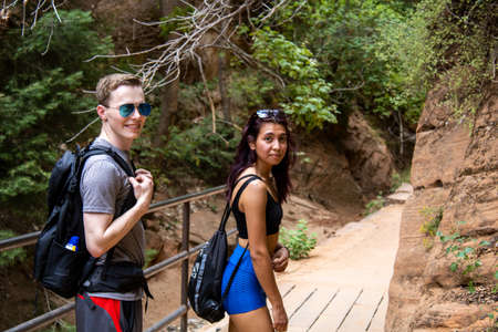 Man woman interracial couple walking down hiking trail looking back at the camera wearing hiker backpacks at Zion National Park side by sideの写真素材
