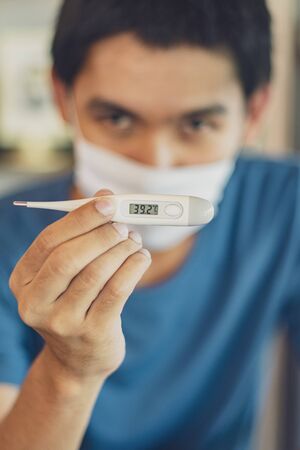Electronic thermometer in the hand man wearing face white mask, against coronavirus. COVID-19 symptoms, high temperature, dry cough.の写真素材