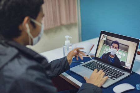 Asian man is working from home during Coronavirus or Covid-19. Wearing a face mask to protect against coronavirus, work at home and using laptop for video conference.  Work from home, Stay home. Social distancing.の写真素材