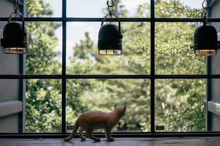 A view of a large square glass window decorated with a lamp. A cat doll stands looking out the window with green leaves in the background.の写真素材