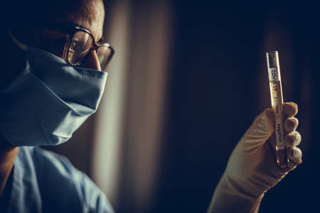 Conceptual photograph of a doctor holding and looking at a test tube while testing samples for presence of coronavirus (COVID-19).の写真素材