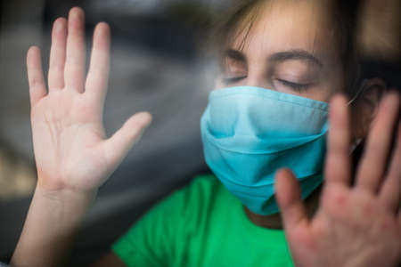 Image of a kid wearing a surgical mask, isolated at home, behind a glass window.の写真素材