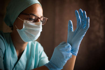 Color portrait of a young female medic arranging her gloves, wearing a mask and glasses,...