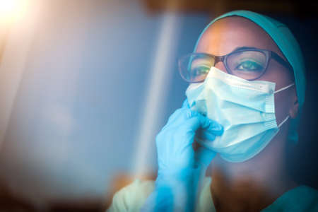 Portrait of a young woman talking on the mobile phone while wearing a mask, indoor, by a window.