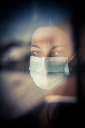 Color image of a woman wearing a surgical mask by a window.の写真素材