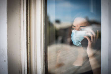 Portrait of a young woman talking on the mobile phone while wearing a mask, indoor, by a window.の写真素材