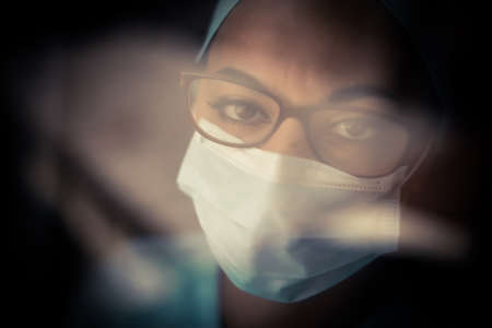 Color portrait of a young female medic by a window, wearing a mask and glasses, illustration for the...