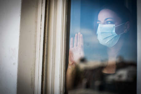Color image of a woman wearing a surgical mask by a window.