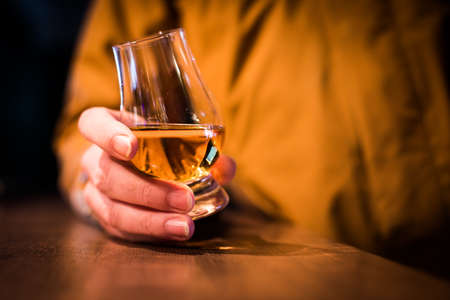 Color close up shot a hand holding a Glencairn whisky glass on a wooden table, with shallow depth of...