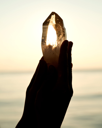 Young woman's hands holding clear quartz point in front of the lake sunriseの写真素材