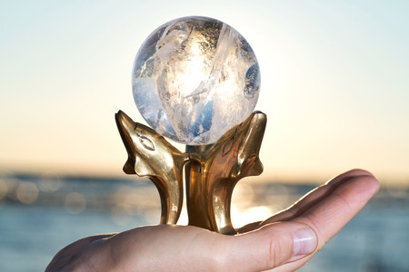 Woman's hand holding Lemurian Clear Quartz Sphere on Dolphins Brass Stand at the sunrise in front of...