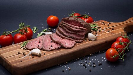 Sliced Grass Fed juicy Corn Roast Beef garnished with Tomatoes, Fresh Rosemary Herb, Garlic and Rainbow Peppercorns on natural wooden cutting board.の写真素材