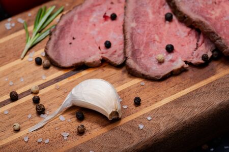Sliced Grass Fed juicy Corn Roast Beef garnished with Fresh Rosemary Herb, Garlic and Rainbow Peppercorns on natural wooden cutting board.の写真素材