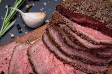 Sliced Grass Fed juicy Corn Roast Beef garnished with Fresh Rosemary Herb, Garlic and Rainbow Peppercorns on natural wooden cutting board.の写真素材