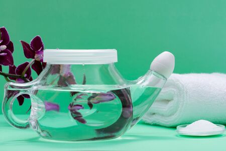 Neti Pot with Soft Comfort Tip, pile of Saline, Purple Orchid Flowers and rolled up White Towels on green background. Sinus wash. Nasal irrigation.の写真素材