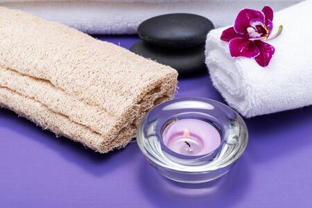 Spa Wellness Concept. Natural Loofah Sponge, rolled up White Towels, Basalt Stones, Orchid and burning Lavender Tea Light Candle on purple background.の写真素材