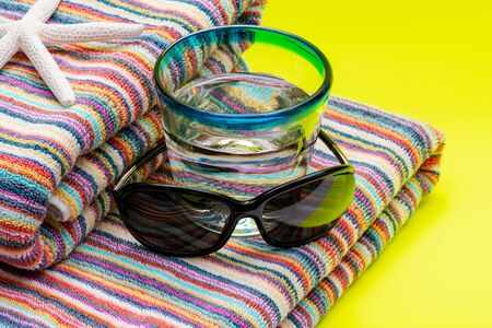 Folded Colorful Striped Organic Cotton Beach Towels, Blue Rim Glass with Water and Black Sunglasses on yellow background as Summer Vacation Concept.の写真素材