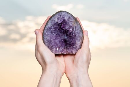 Woman's hand holding partially polished Heart shaped Amethyst geode specimen from Brazil at sunrise in front of the lake.の写真素材