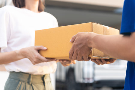 Close up delivery man hands wearing blue uniform send a cardboard box to customer in front of the customer's house with sunset flare background.の写真素材