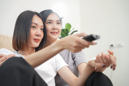 Affection LGBT concept. Happy diverse beautiful asian female lesbian couple sitting on sofa at home hugging and laughing as they watch evening TV together in slow motion shot. Spending quality time.の写真素材