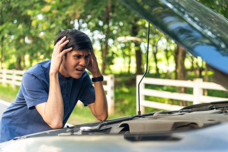 Asian man repairing a broken car by the road. Man having trouble with his broken car on the highway roadside. Man looking under the car hood.の写真素材