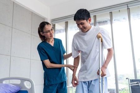 Beautiful asian woman physiotherapist assists Asian man patient in walking exercises using crutches in physical therapy clinic.の写真素材