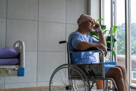 Asian man cancer patient sitting alone in wheelchair looking outside window in hospital ward with...