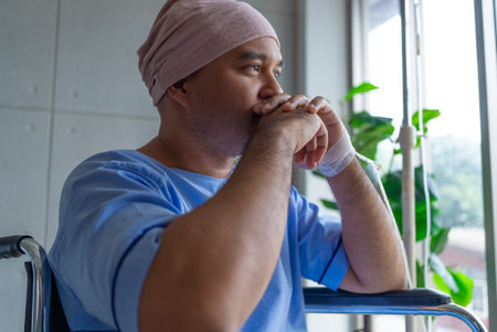 Asian man cancer patient sitting alone in wheelchair looking outside window in hospital ward with...