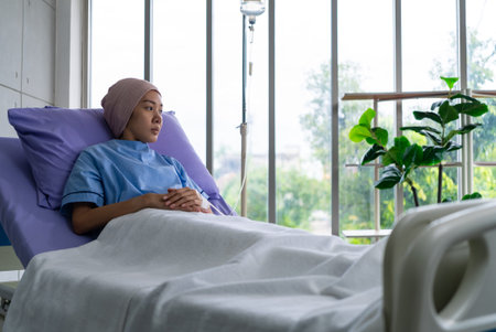 Asian woman cancer patient sitting alone on a hospital bed receiving saline and looking outside...
