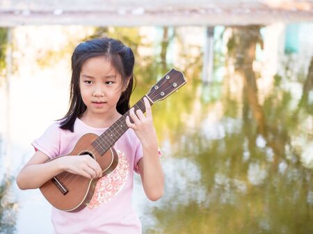Little asian child girl play the ukulele, in the garden near the pondの写真素材