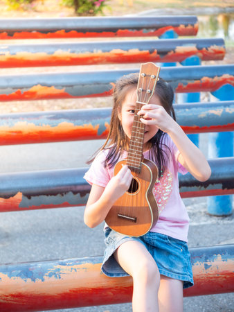 Little asian child girl play the ukulele, in the garden on the Steel pipe, practice to play