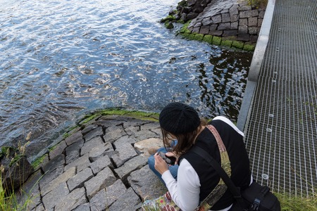 A young woman sitting by the water of the Kiel Canal and plays with her smartphone の写真素材