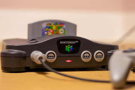 A Nintendo 64 Console With the Power Indicator Light On and Super Mario 64 in the Cartridge Slot and a Controller Plugged Into Itのeditorial素材