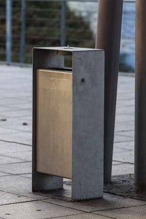 An outdoor metal waste bin stands near a pillar on a square...