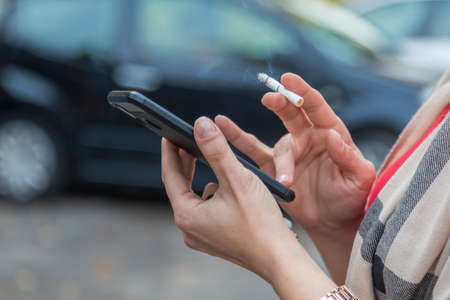 Female hands holding a mobile phone and having a lit cigarette between them, which is smoking and a car in the backgroundの写真素材