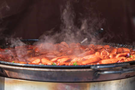 Sausages in their own juice on a large round pan from which steam comes, placed on the counter and intended for sale.の写真素材