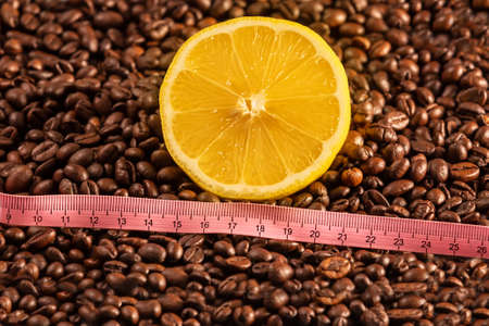 Half of a yellow lemon lies on a layer of coffee beans. An inch is placed in front of the lemon.